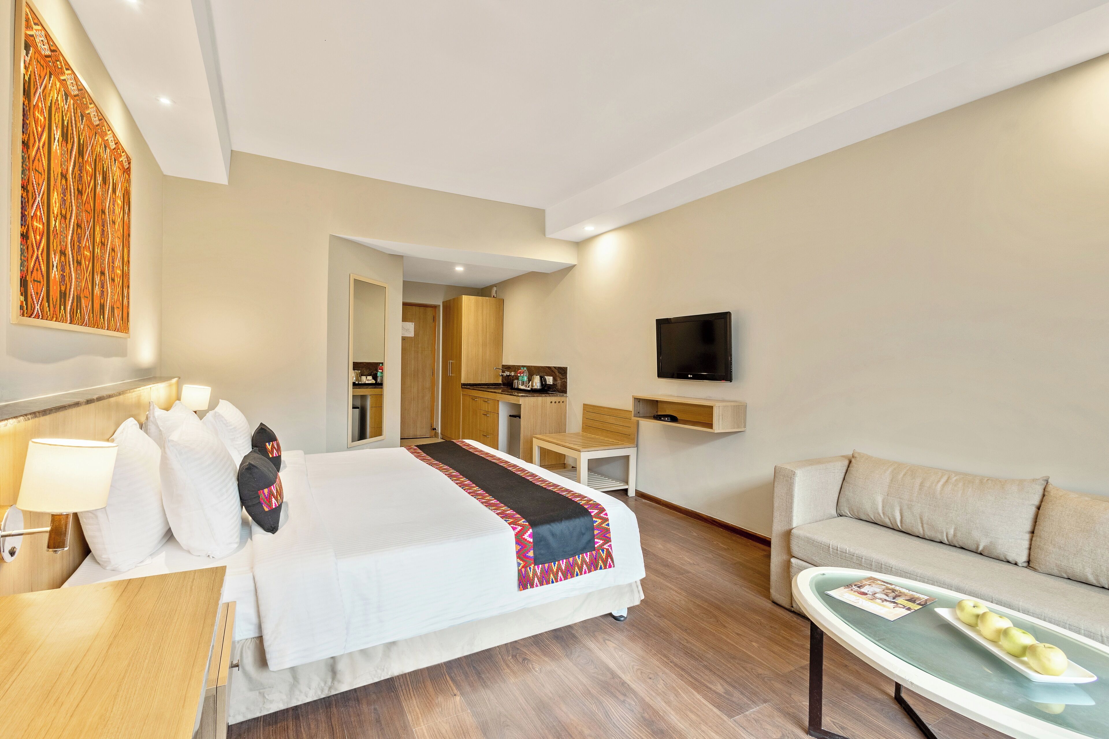 A hotel room with a bed, a desk, a sofa, a TV, and a dining table with apples.