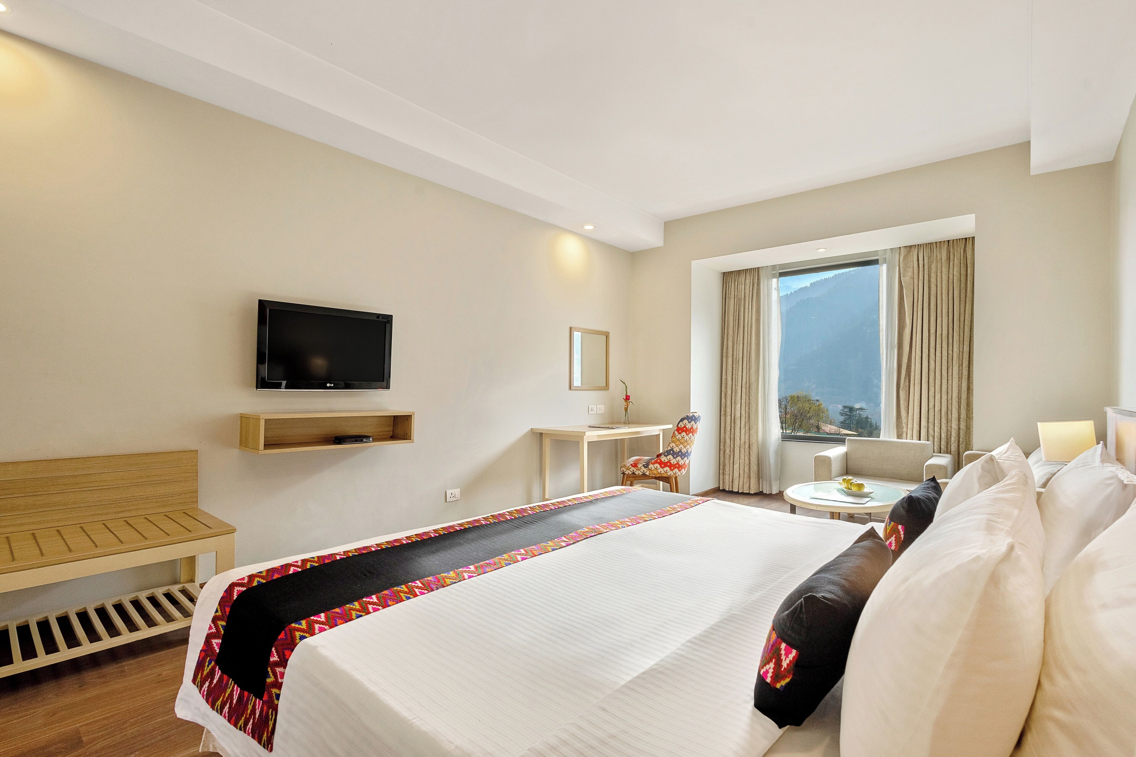 Habitación de hotel con cama, televisión, escritorio, silla, mesita y ventana con cortinas.