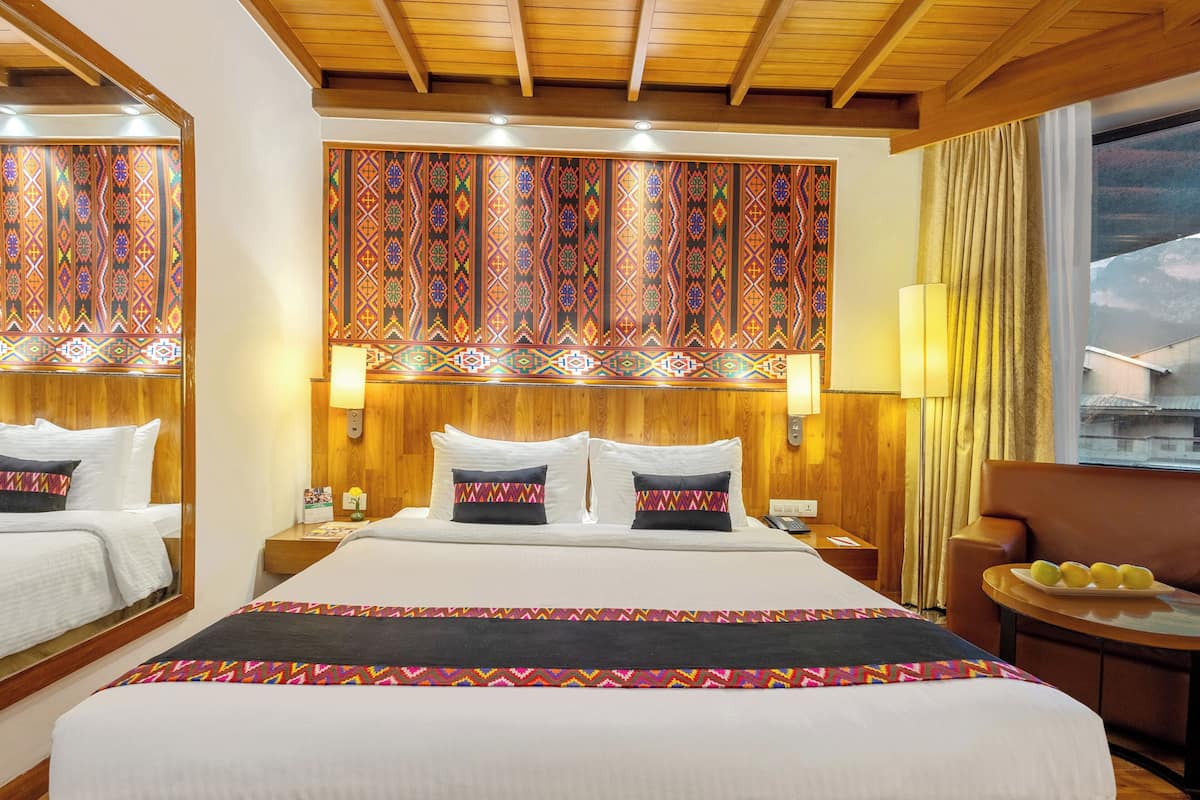 Ein Hotelzimmer mit einem großen Bett, einem Holzkopfende und einer dekorativen Wandverkleidung. Es gibt einen kleinen Tisch mit einer Fruchtschale und einen Stuhl.
