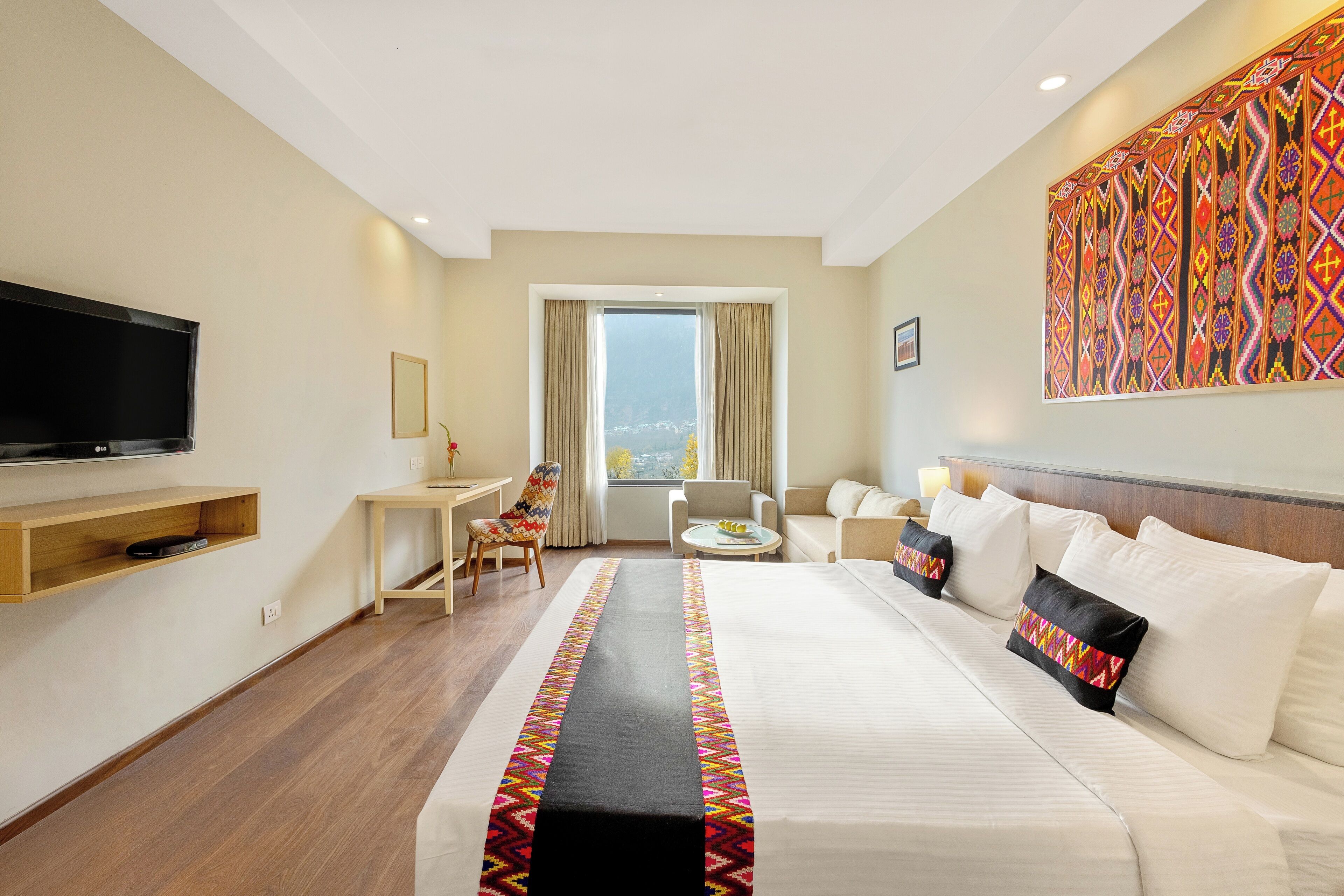 Ein Hotelzimmer mit einem großen Bett, einem an der Wand montierten Fernseher, einem Schreibtisch, einem Sessel und Blick auf die Stadt durch das Fenster.