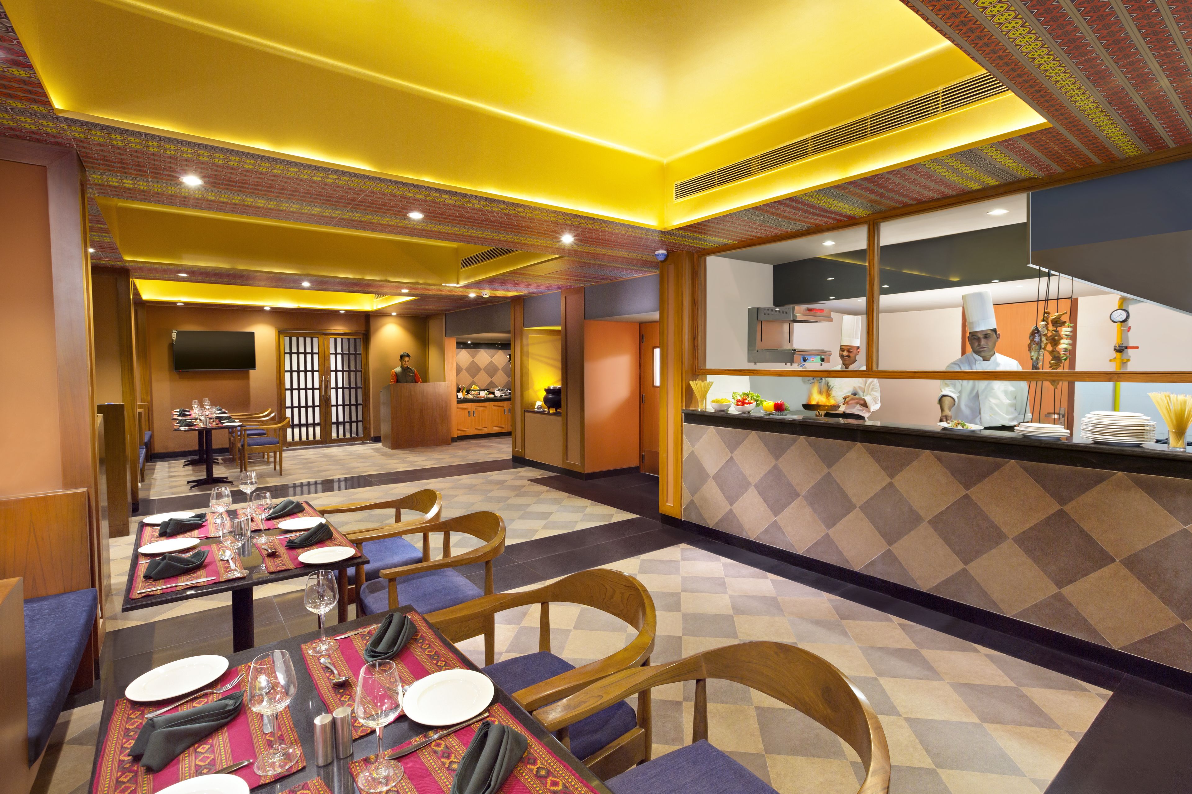 A restaurant with tables set for dining, a kitchen area, and a seating area with cushioned chairs.
