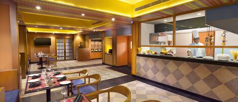 A restaurant with tables set for dining, a kitchen area, and a seating area with cushioned chairs.