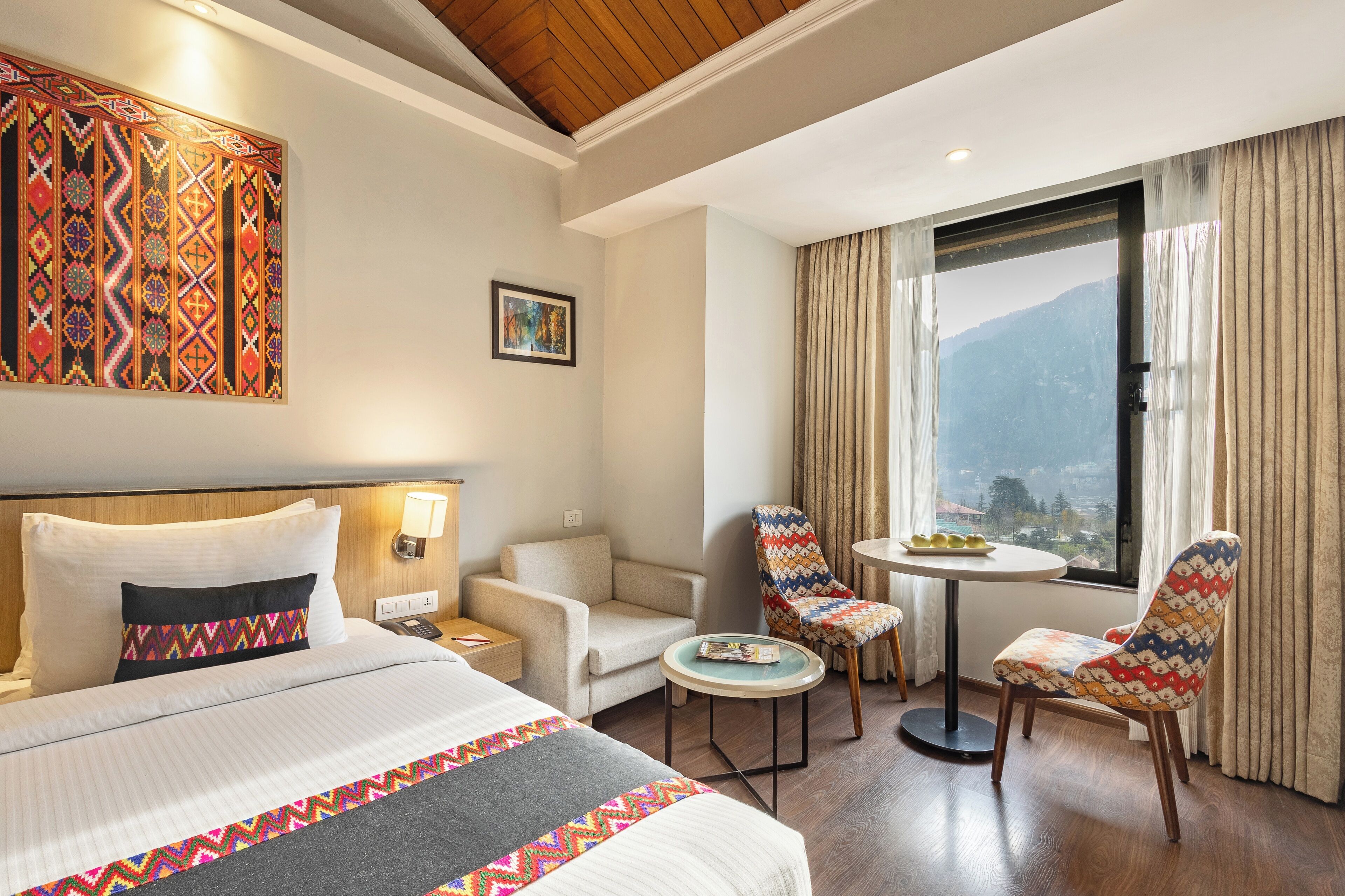 A hotel room with a bed, two chairs, a small table, a painting, and a window with a mountain view.