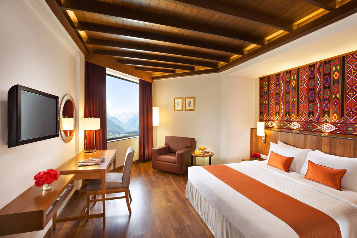 Ein Hotelzimmer mit Holzbalkendecke, einem Bett mit orangefarbener Bettwäsche, einem Schreibtisch mit Stuhl, einem Fernseher und einem Fenster mit Bergblick.