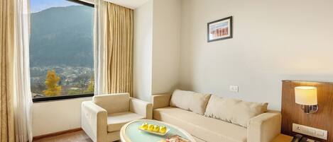 A hotel room with a sofa, armchair, and a small table with a magazine. There is a window with curtains, a wall-mounted lamp, and a framed picture.