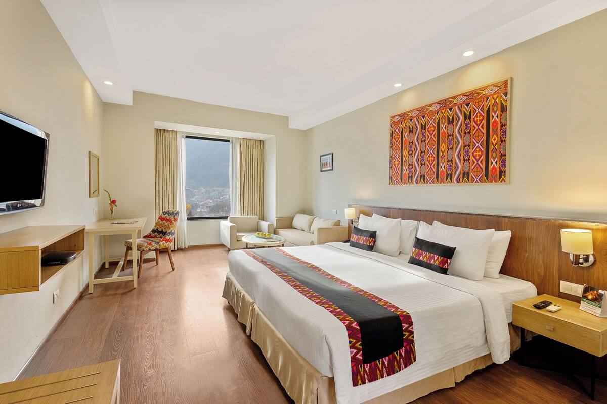 Ein Hotelzimmer mit einem großen Bett, einem Schreibtisch, einem Sessel, einem Fernseher und einem Fenster mit Blick auf die Stadt.