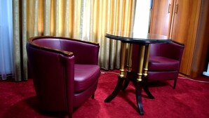 A room with two purple armchairs and a glass-top table with gold legs, set against a red carpet and a backdrop of curtains. - Aristocrat Hotel (Skopje)