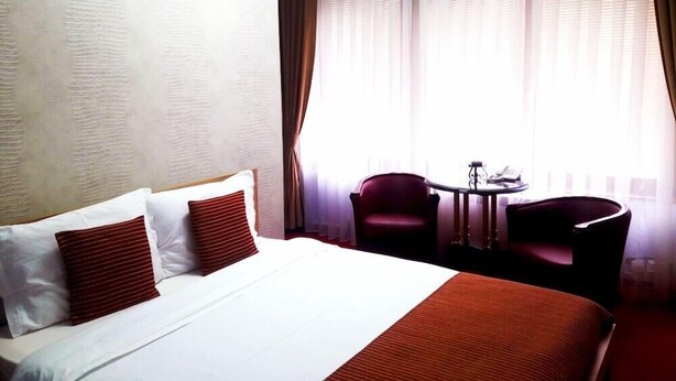 A hotel room with a large bed, two red pillows, a small table with a chair, and a window with sheer curtains. - Aristocrat Hotel (Skopje)