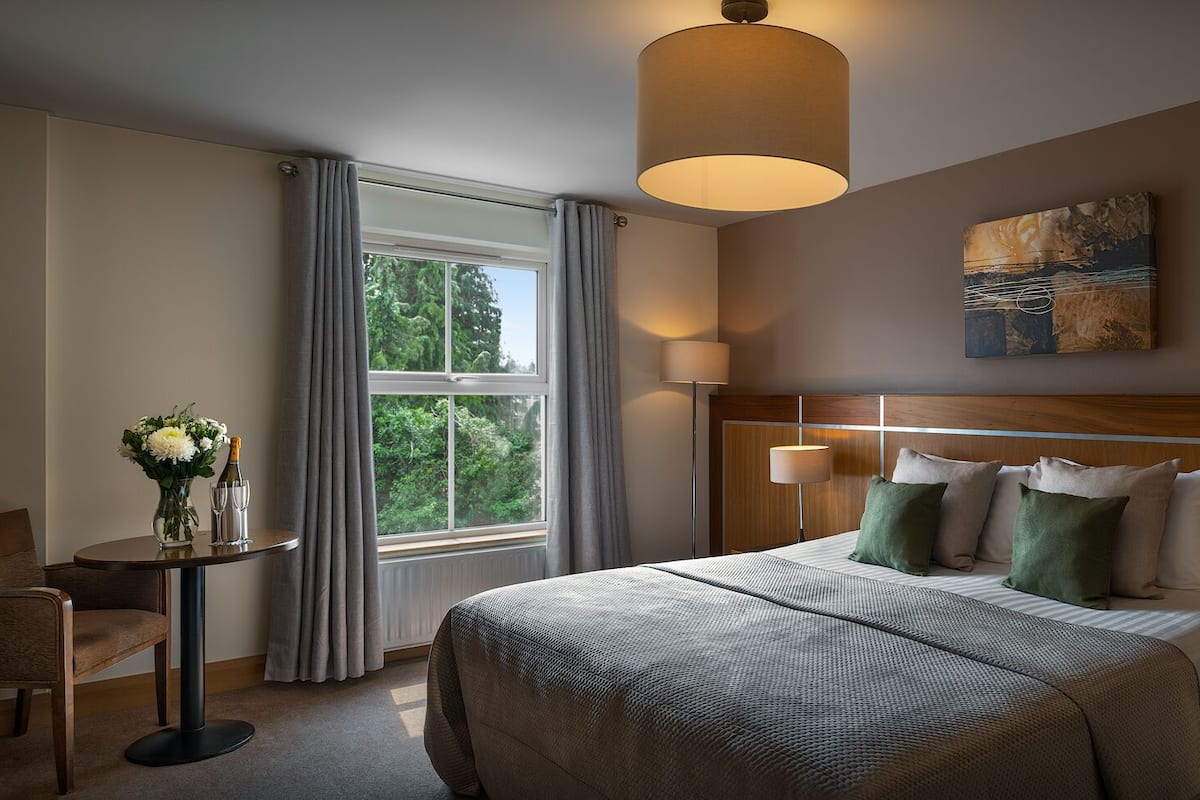 Une chambre d’hôtel avec un grand lit, deux tables de chevet avec des lampes, une petite table avec un vase et des coupes à champagne, une chaise et une fenêtre avec des rideaux.