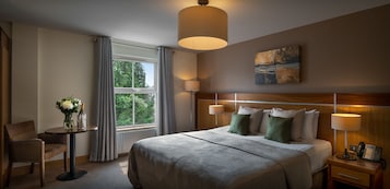Una habitación de hotel con una cama grande, dos mesitas de noche con lámparas, una pequeña mesa con un florero y copas de champán, una silla y una ventana con cortinas.