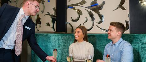 A restaurant setting with a table set for three people, a waiter serving drinks, and a decorative wall with abstract patterns.