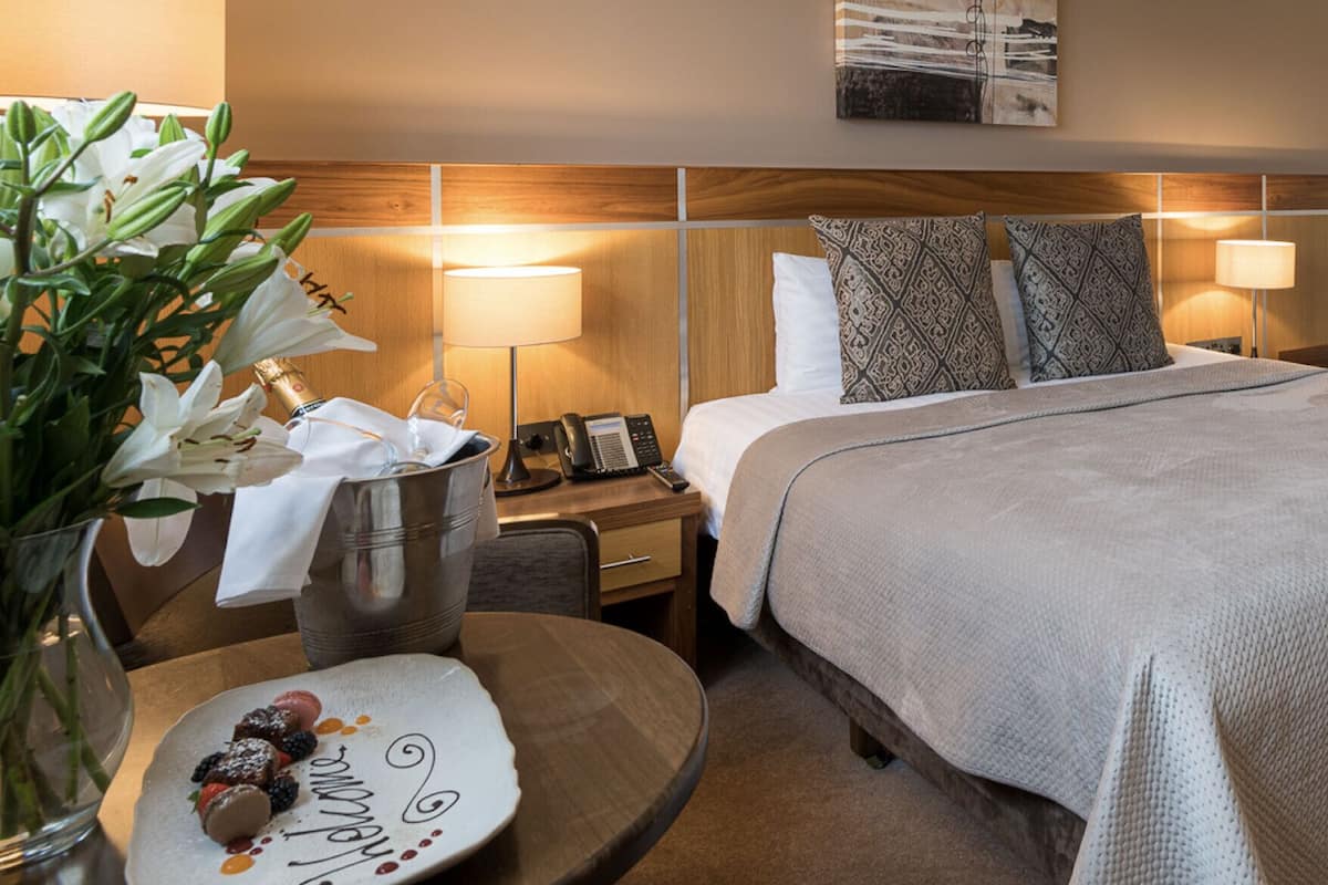 Une chambre d’hôtel avec un lit, des lampes de chevet, une table de chevet avec un téléphone, un vase de fleurs et une assiette avec de la nourriture.