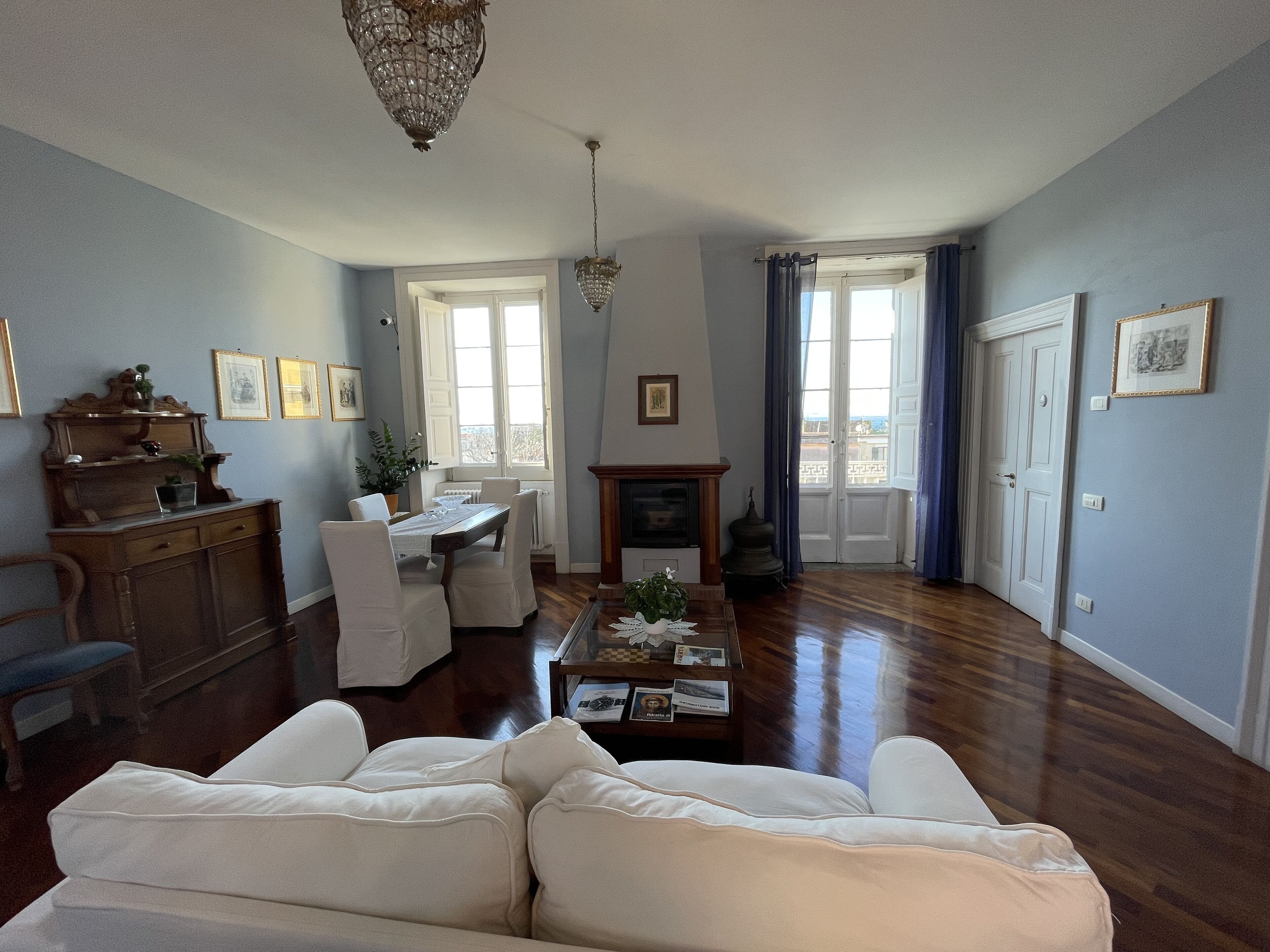 A spacious living room with a wooden floor, a white sofa, a dining table with chairs, a chandelier, and a fireplace.
