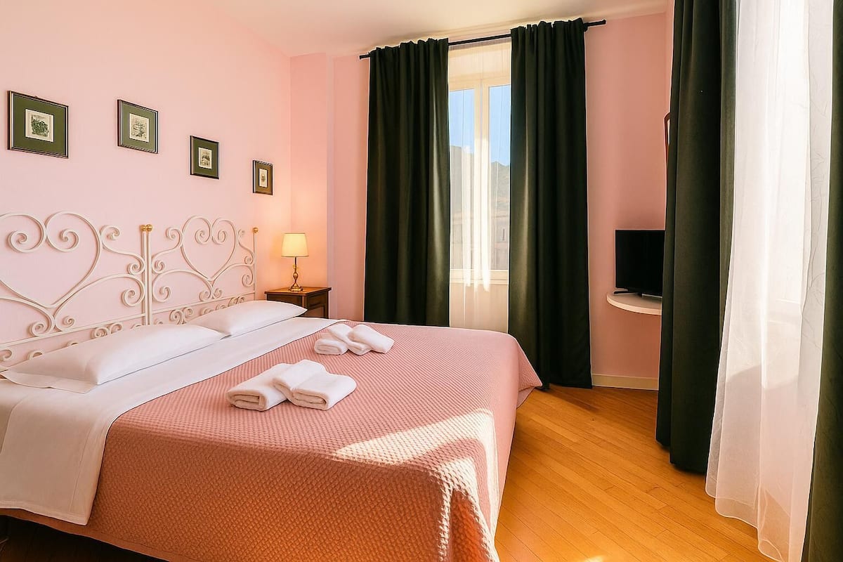 Een net opgemaakt bed met witte lakens en roze beddengoed, een bedlampje, een flatscreen televisie op een standaard en een raam met donkere gordijnen.