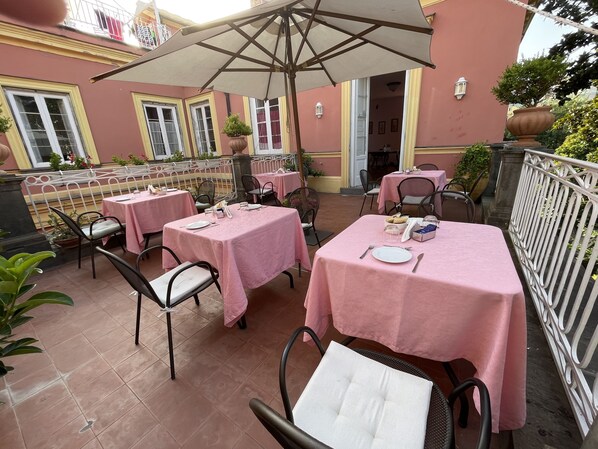 An outdoor patio with tables and chairs, covered by umbrellas, and a building with yellow trim in the background. - The Secret Garden Relais (Piano di Sorrento)