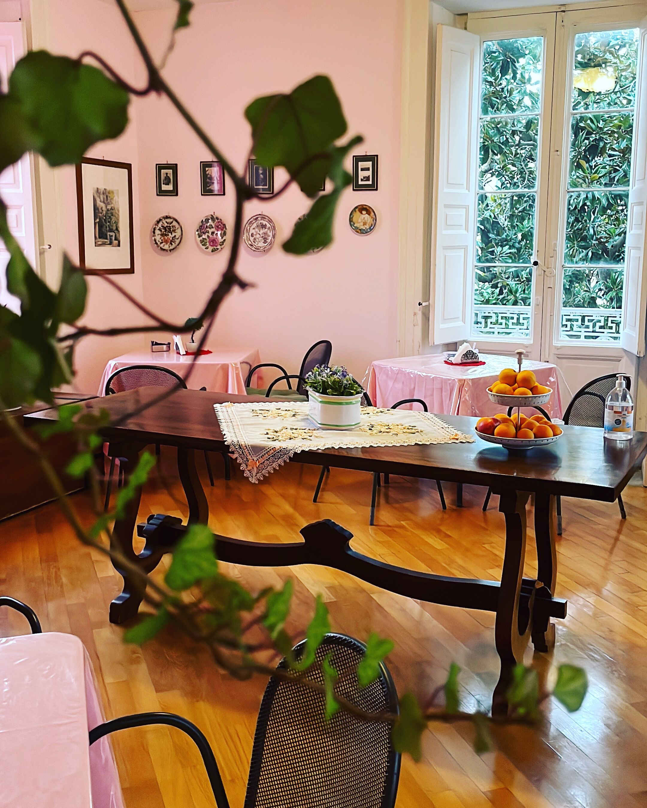A dining area with a wooden table, chairs, and a pink tablecloth. There are framed pictures on the wall and a window with a view of greenery.