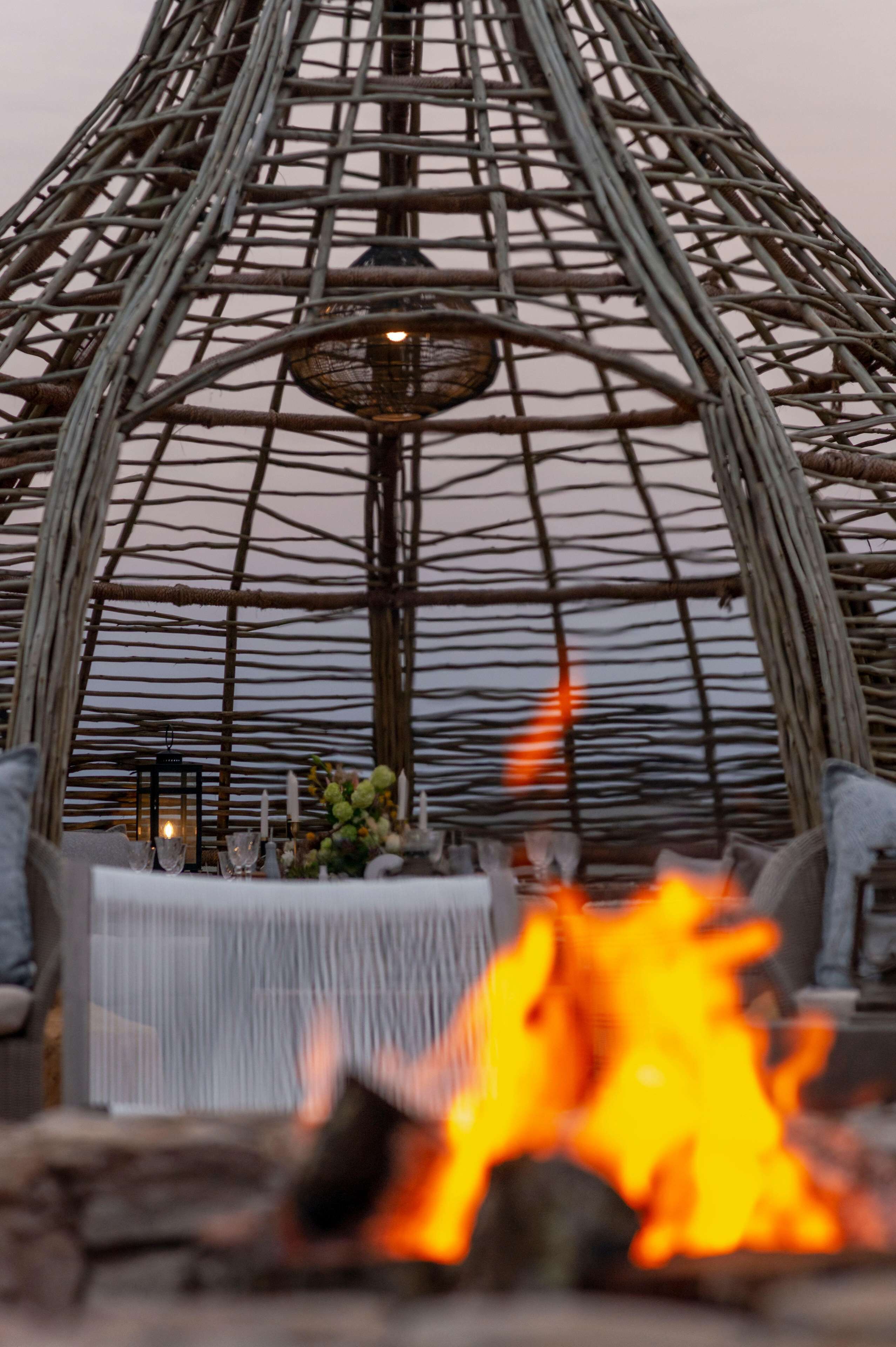 Un espace repas avec une structure en osier au-dessus, une table dressée avec des verres et une bougie, et un feu au premier plan.