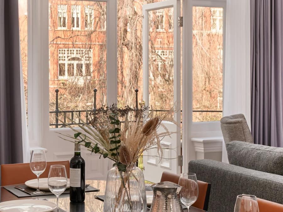 Mesa de jantar posta para quatro com vista para um edifício de tijolos vermelhos através de uma grande janela arqueada.