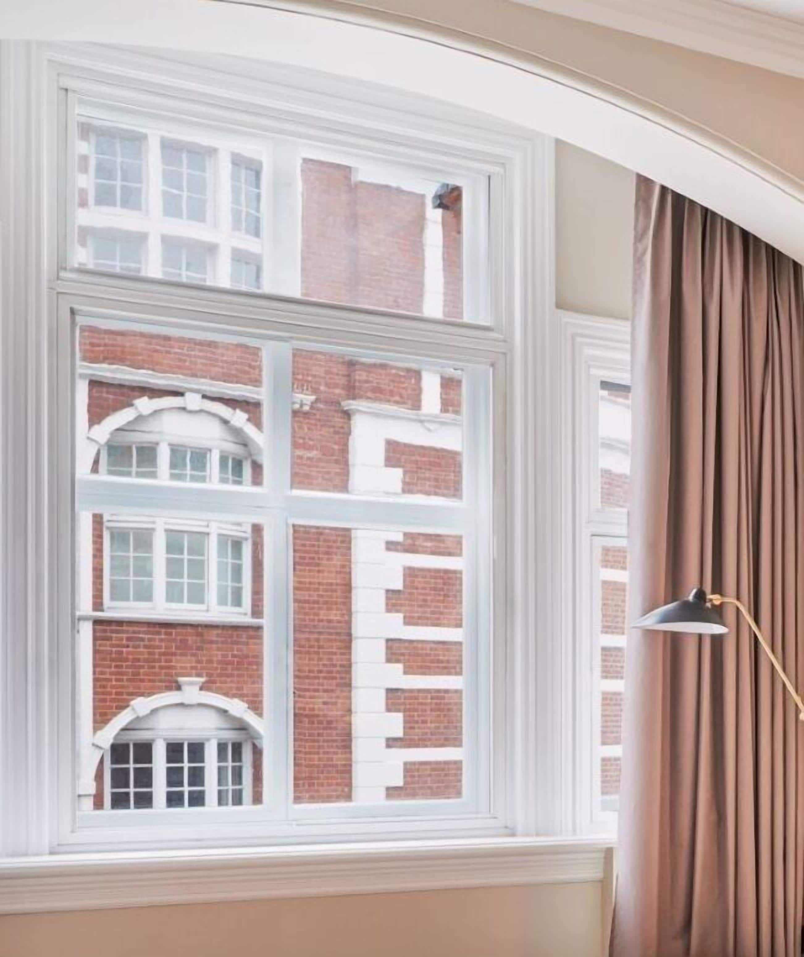 A room with a large window overlooking a brick building, a floor lamp, and a curtain.