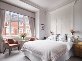 Ein Schlafzimmer mit einem großen Bett, einem Sessel, einem kleinen Tisch mit Blumenvase und Blick auf Gebäude durch ein Fenster.