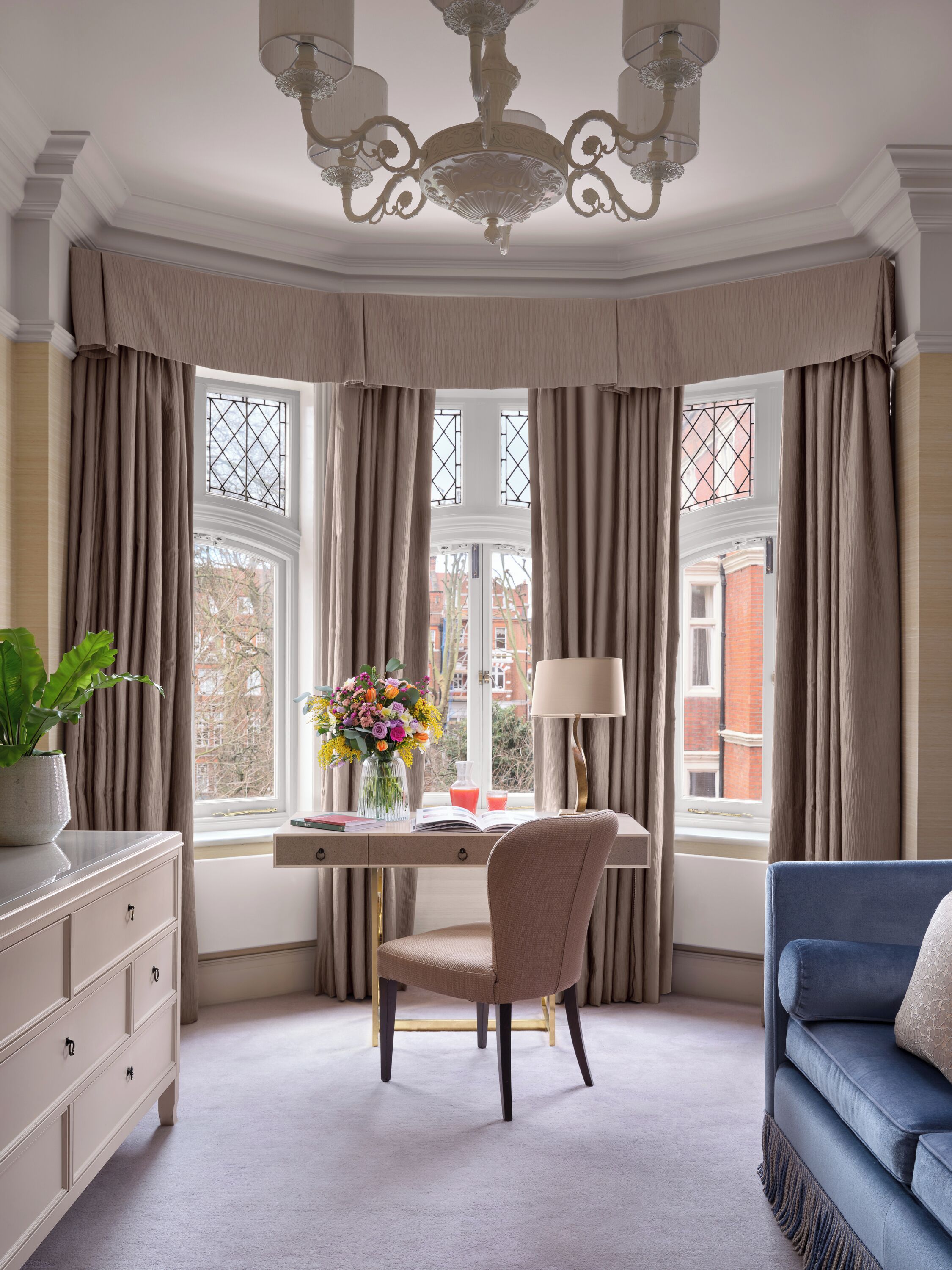 A corner room with a desk, chair, and a blue sofa. There is a window with curtains, a chandelier, and a vase with flowers on the desk.