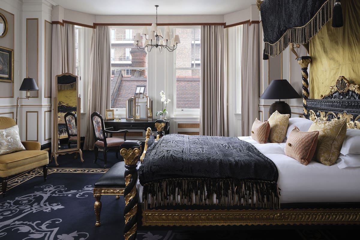 A luxurious bedroom with a large bed, a desk, and a chair. There is a view of the outside through large windows.