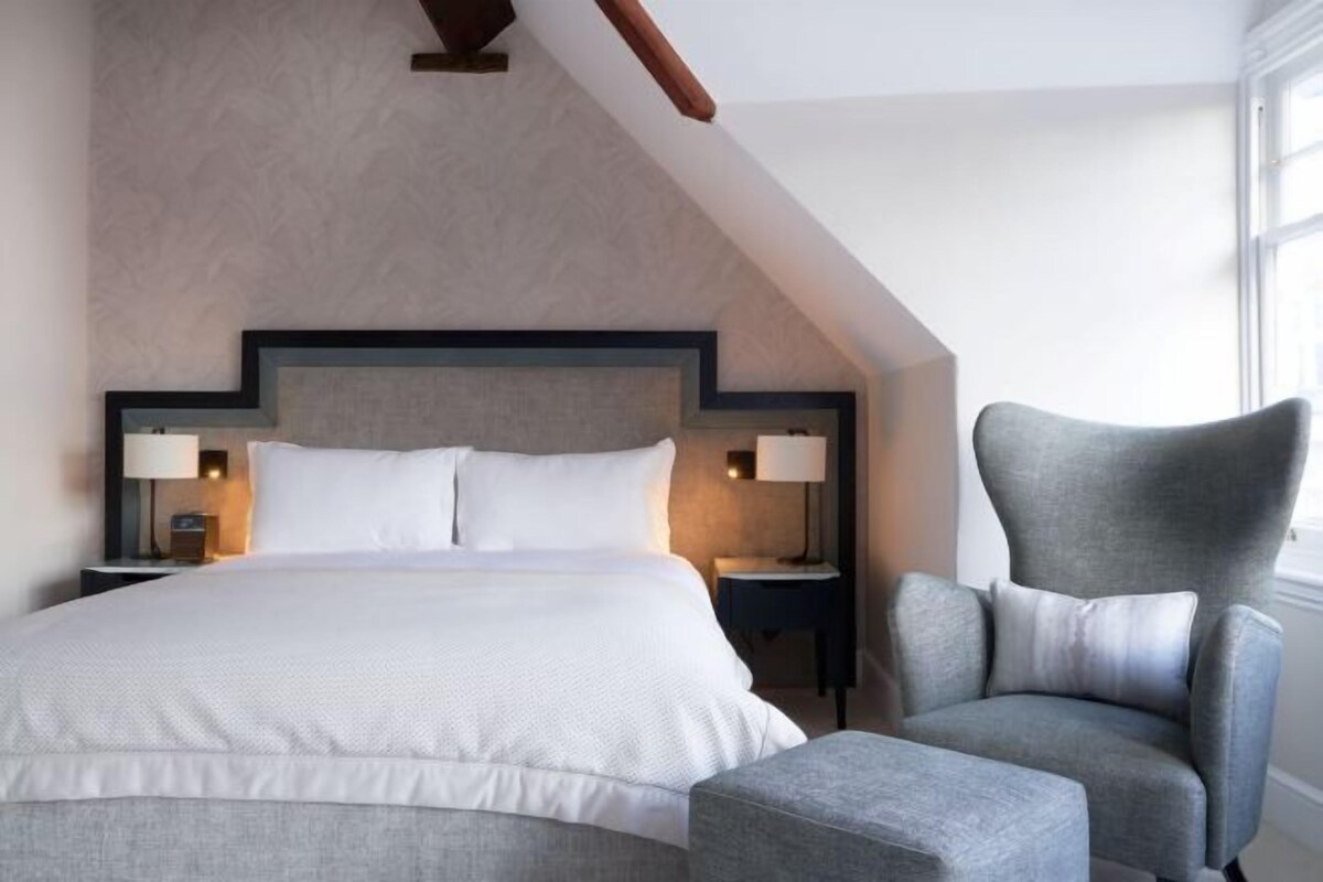 A hotel room with a large bed, two bedside tables with lamps, a grey armchair, and a window.