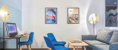 A modern room with a desk, two blue chairs, a small table, a sofa, and two framed pictures on the wall.