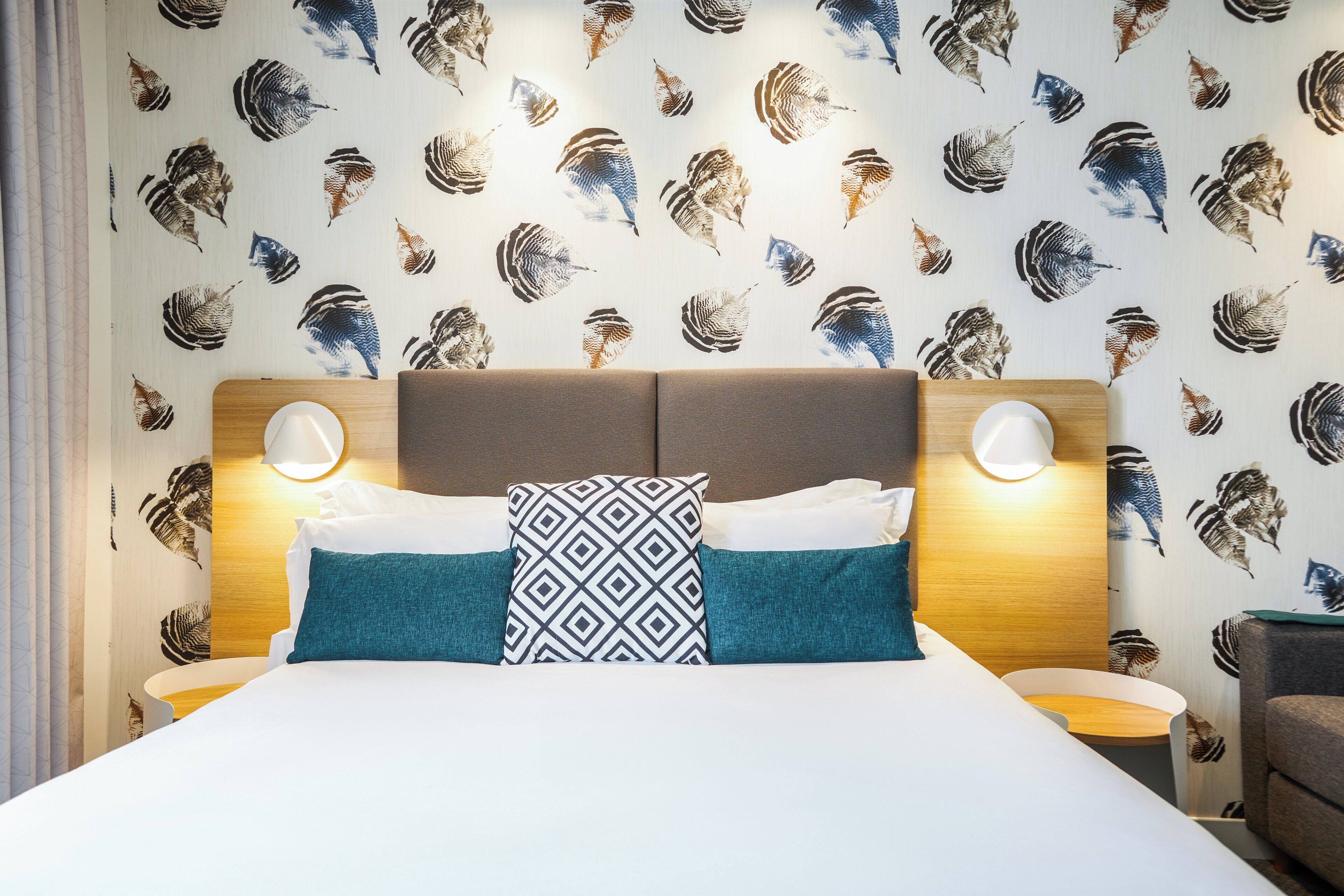 Una habitación de hotel con una cama, dos almohadas, cabecero y lámparas de noche, y una pared con estampado de pájaros.