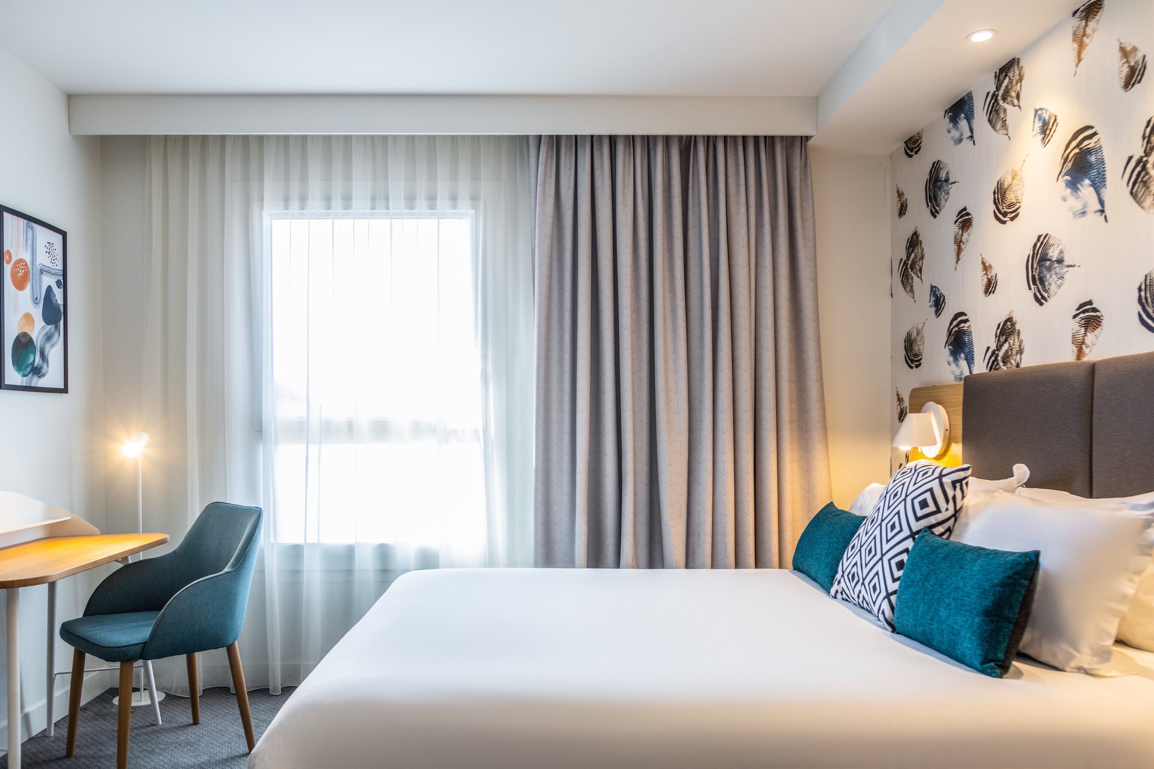 Habitación de hotel con cama, escritorio, silla y un amplio ventanal con cortinas.