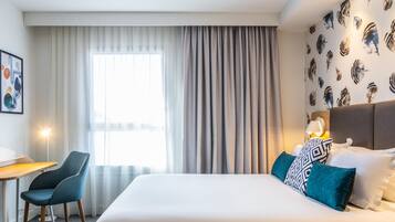 Habitación de hotel con cama, escritorio, silla y un amplio ventanal con cortinas.