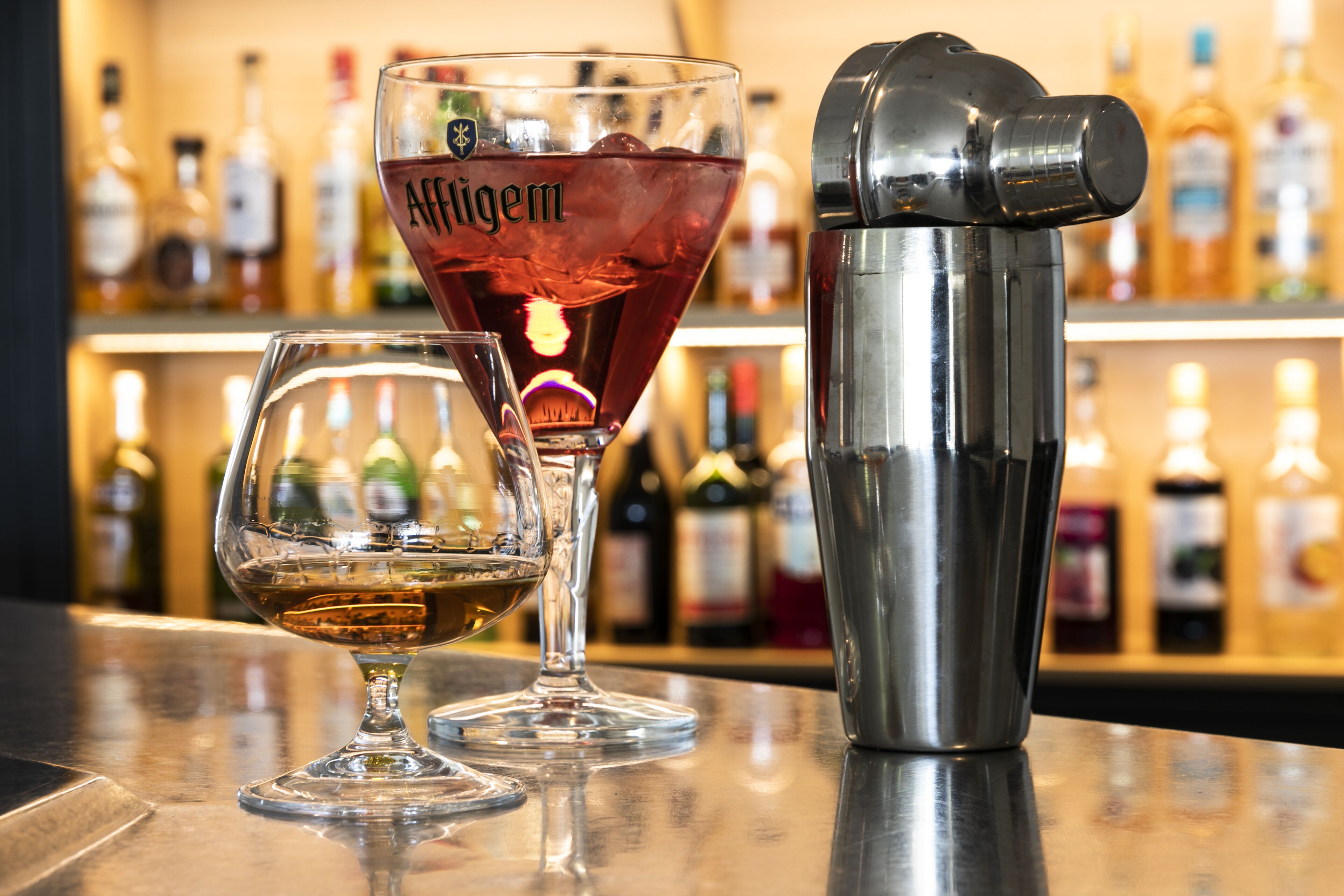 Deux verres contenant des boissons alcoolisées, dont l’un porte une étiquette, et un shaker à cocktails, le tout posé sur le comptoir d’un bar.
