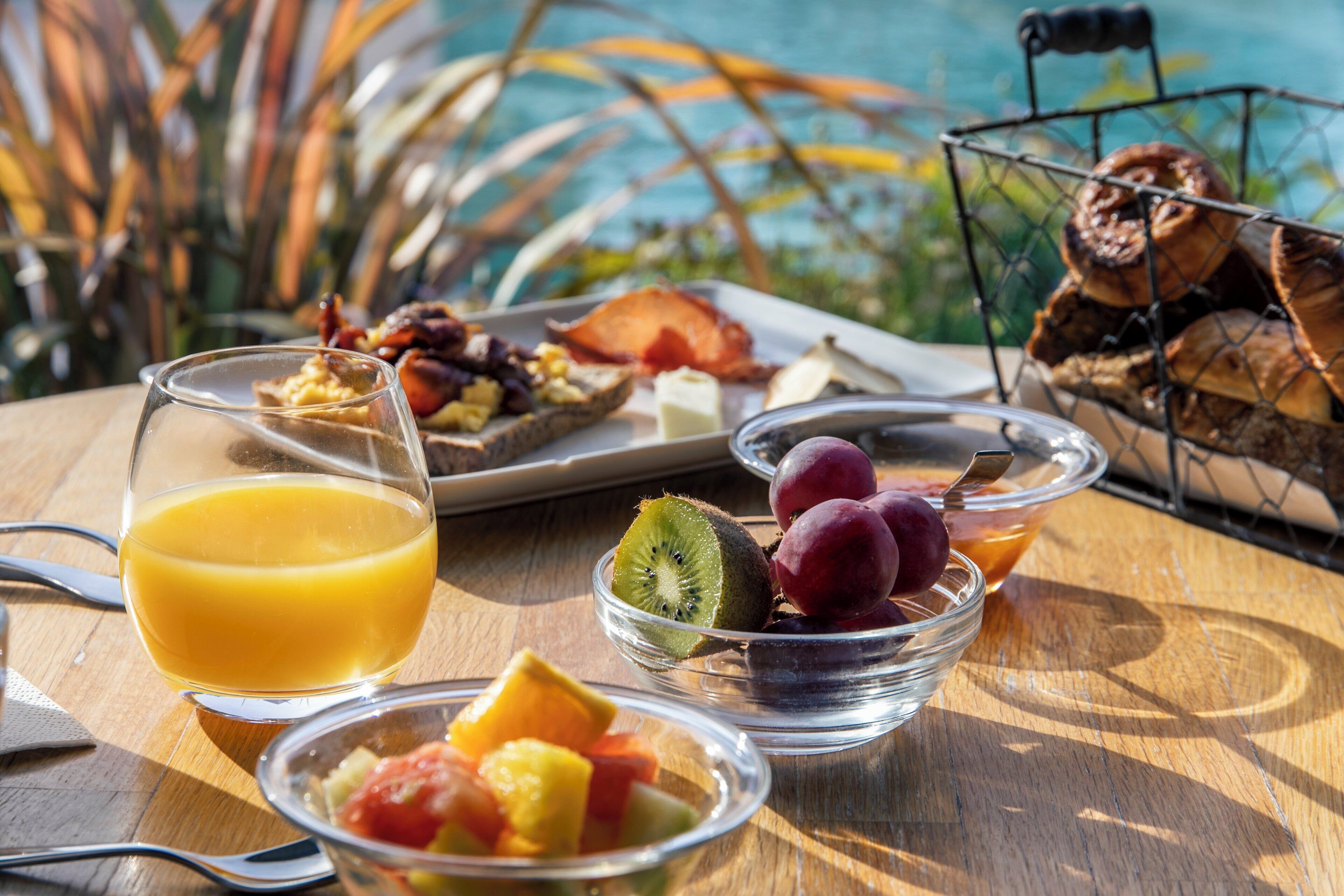 A breakfast table with a glass of orange juice, a plate of eggs, bacon, and toast, a bowl of fruit, and a basket of pastries.