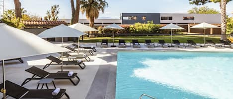 Una piscina con tumbonas y sombrillas, rodeada de árboles y un edificio.