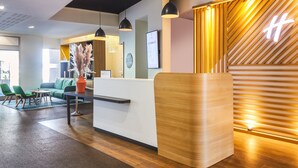 A reception area with a wooden reception desk, green chairs, a sofa, and pendant lighting. - Holiday Inn Perpignan by IHG (Perpignan)
