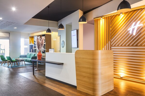 A reception area with a wooden reception desk, green chairs, a sofa, and pendant lighting. - Holiday Inn Perpignan by IHG (Perpignan)