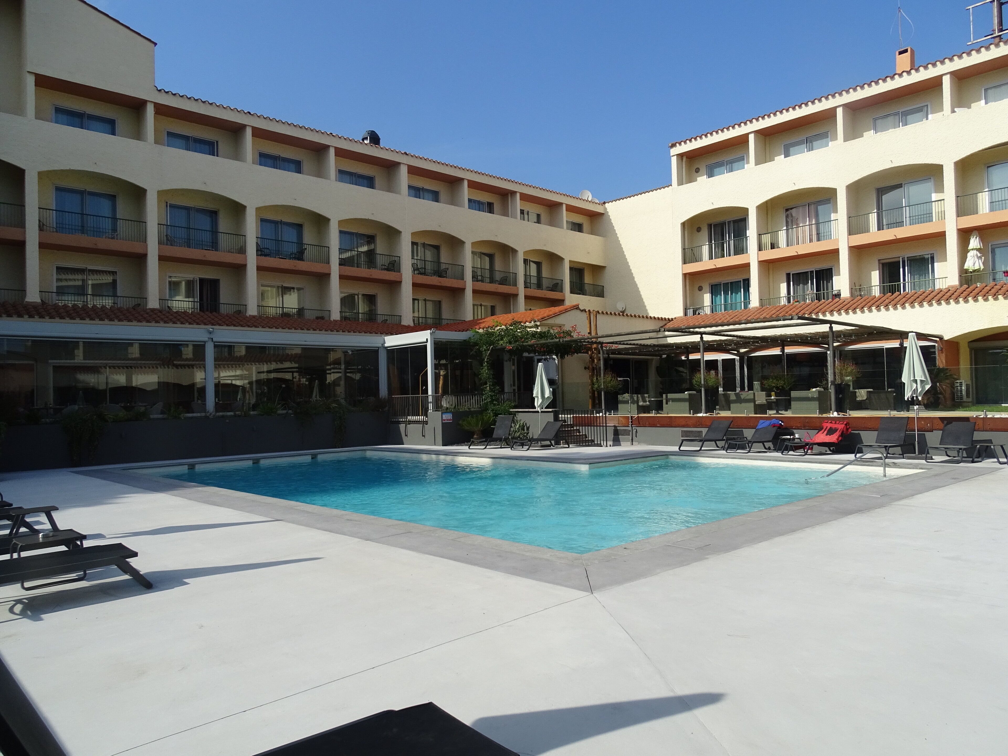 A hotel pool area with lounge chairs and umbrellas, surrounded by a multi-story building with balconies.