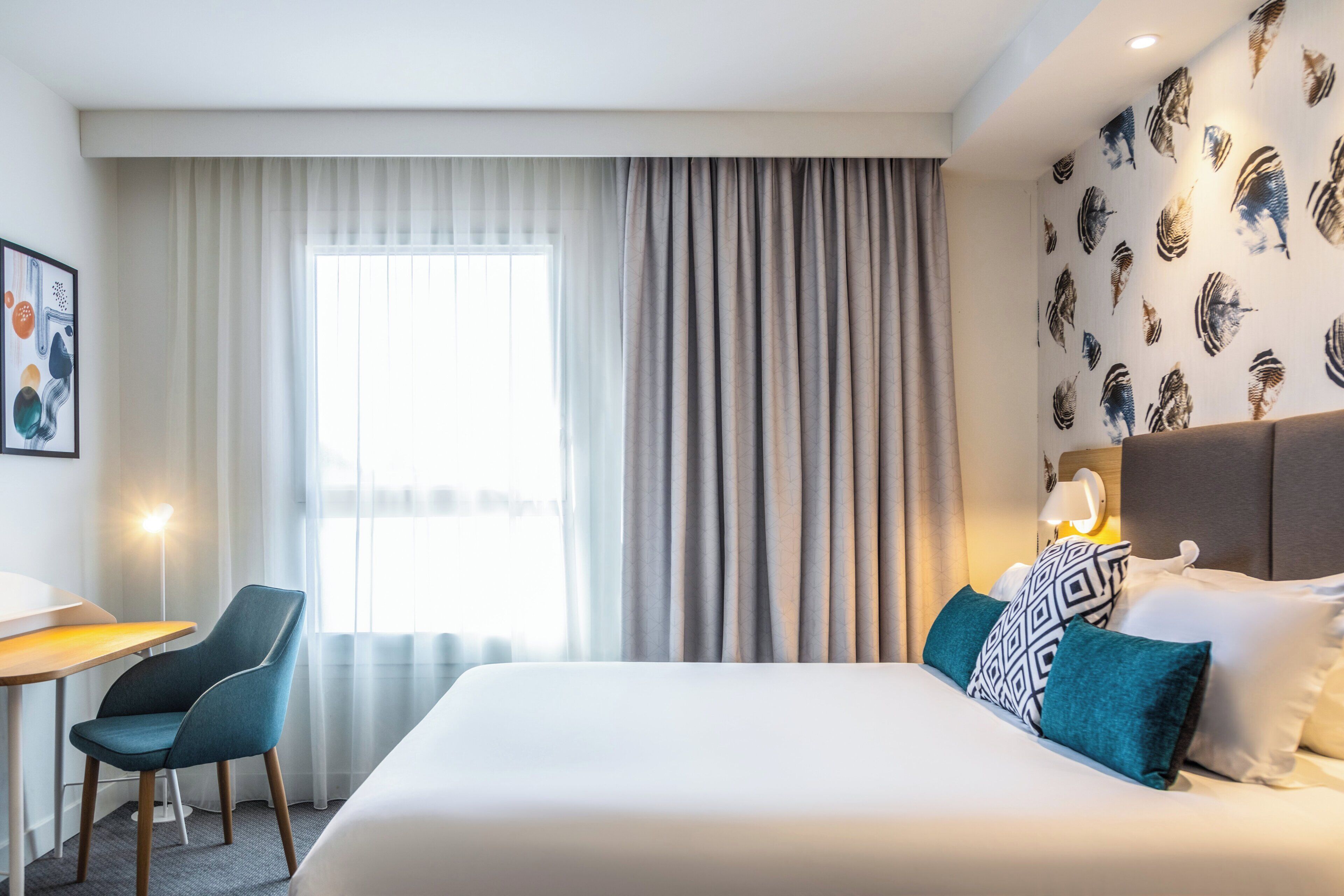Ett hotellrum med en säng, ett skrivbord, en stol och ett fönster med gardiner.