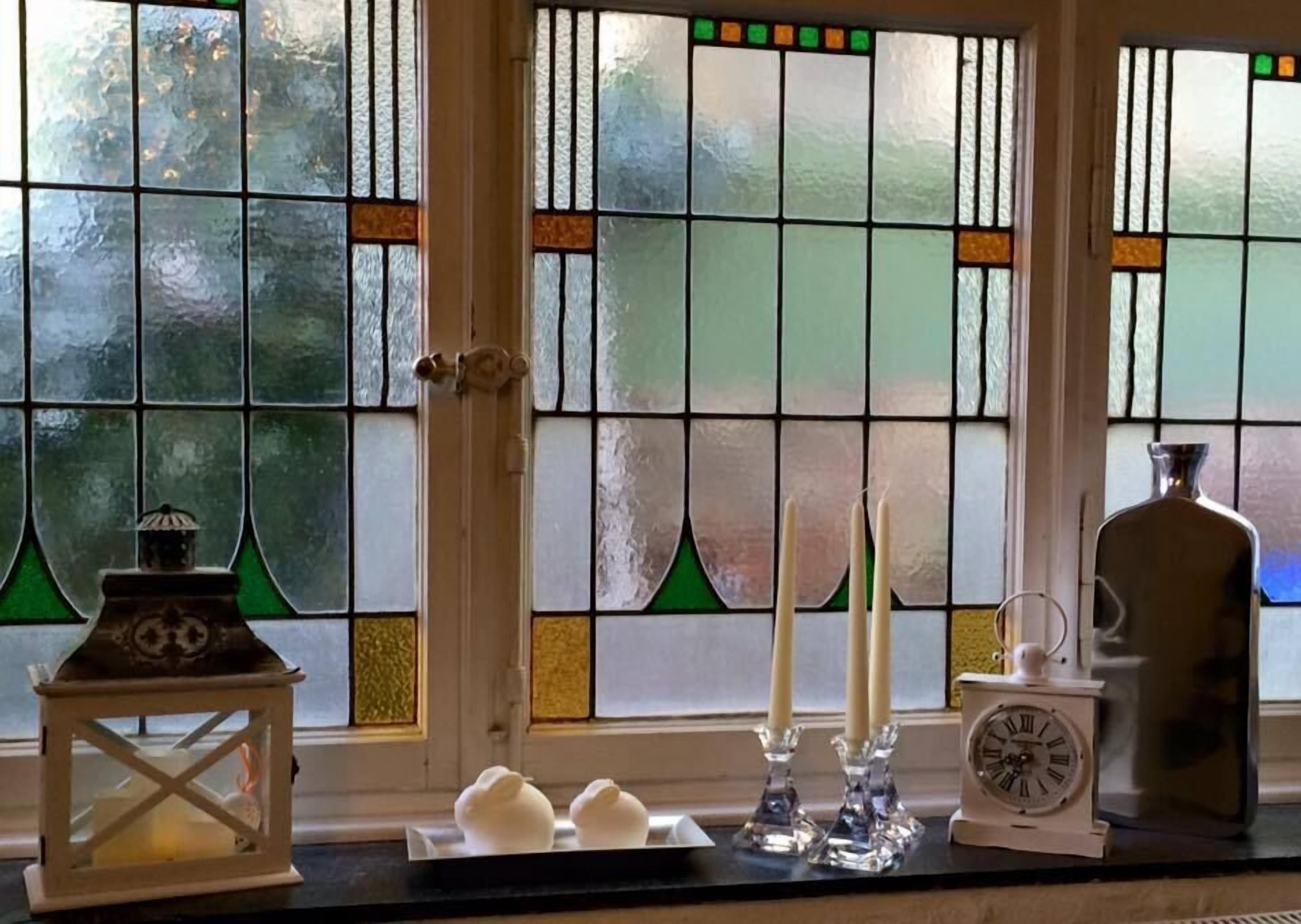 Une pièce avec une fenêtre ornée de vitraux, une lanterne blanche, deux bougies et une horloge posée sur le rebord de la fenêtre.