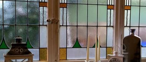 Une pièce avec une fenêtre ornée de vitraux, une lanterne blanche, deux bougies et une horloge posée sur le rebord de la fenêtre.
