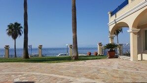 Une terrasse dallée agrémentée de palmiers, avec vue sur la mer, et un bâtiment orné d’arcades.