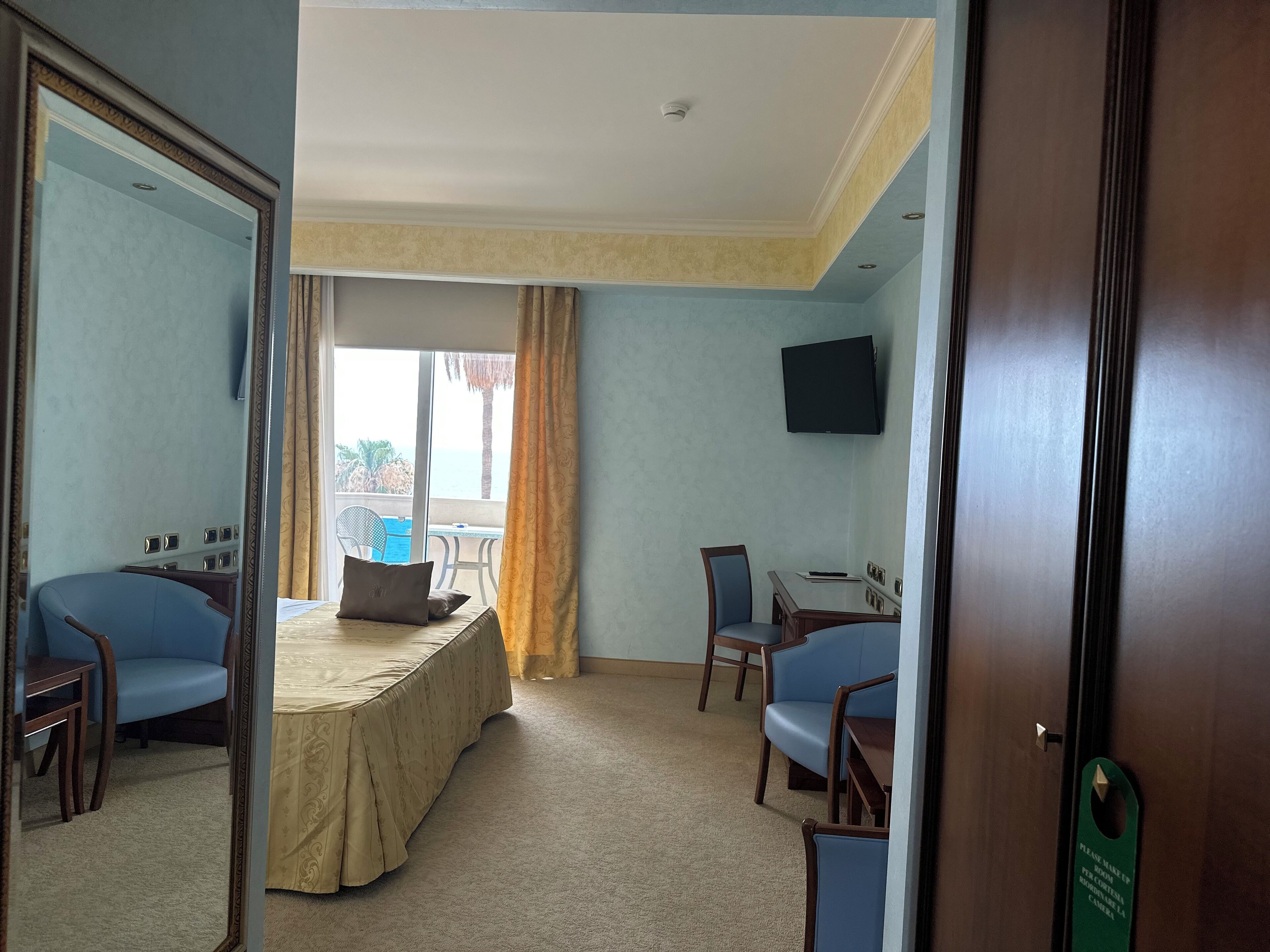 A hotel room with a bed, two blue chairs, a desk, a TV, and a view of the sea through a large window.
