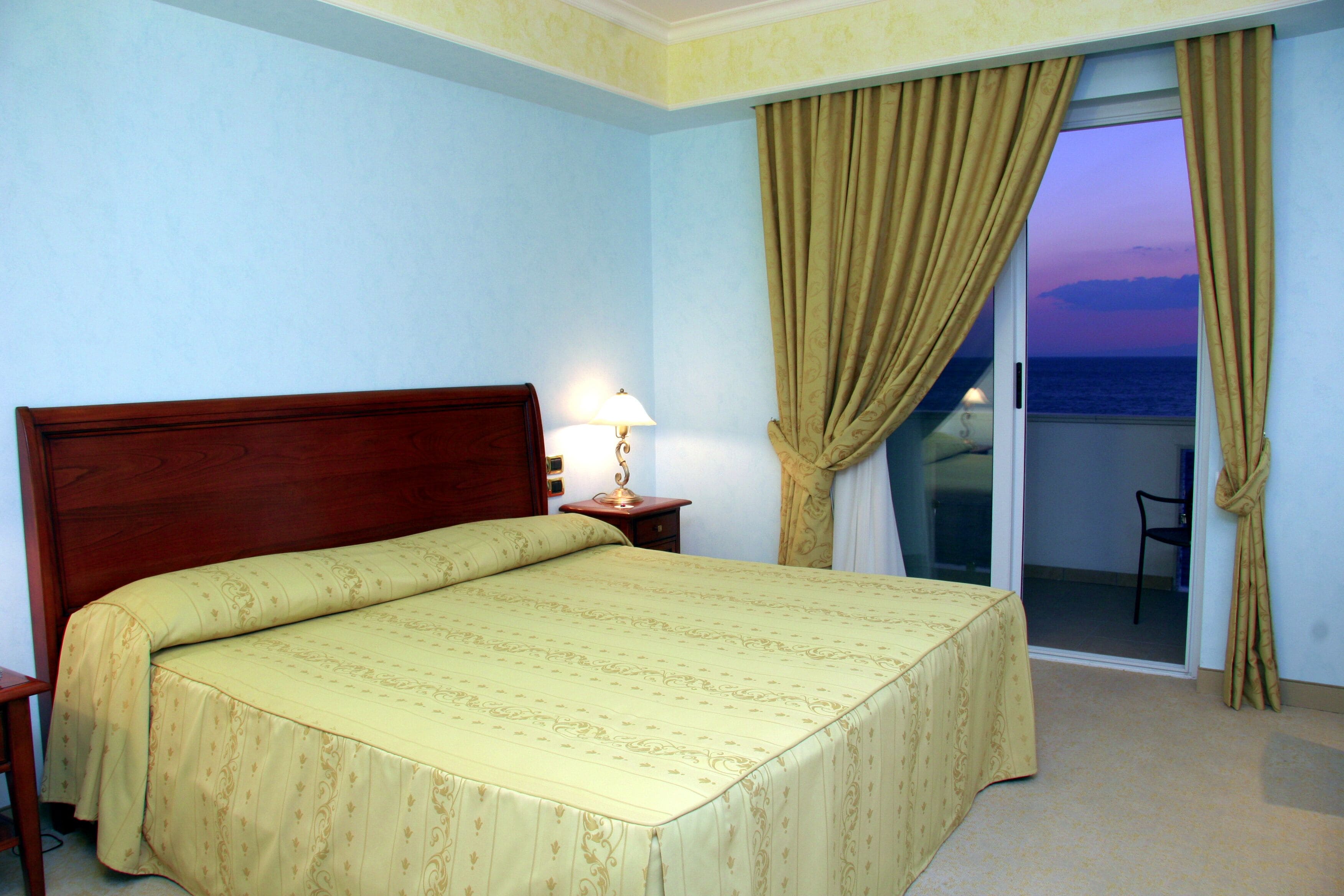 Una camera d'hotel con un letto, un comodino, una lampada e vista mare attraverso una porta.