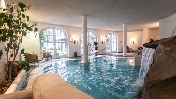 Una piscina interior con caída de agua, ventanas amplias y sillones de descanso.