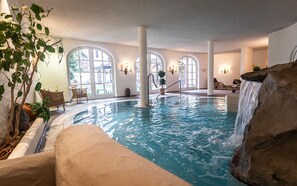 Una piscina interior con caída de agua, ventanas amplias y sillones de descanso.