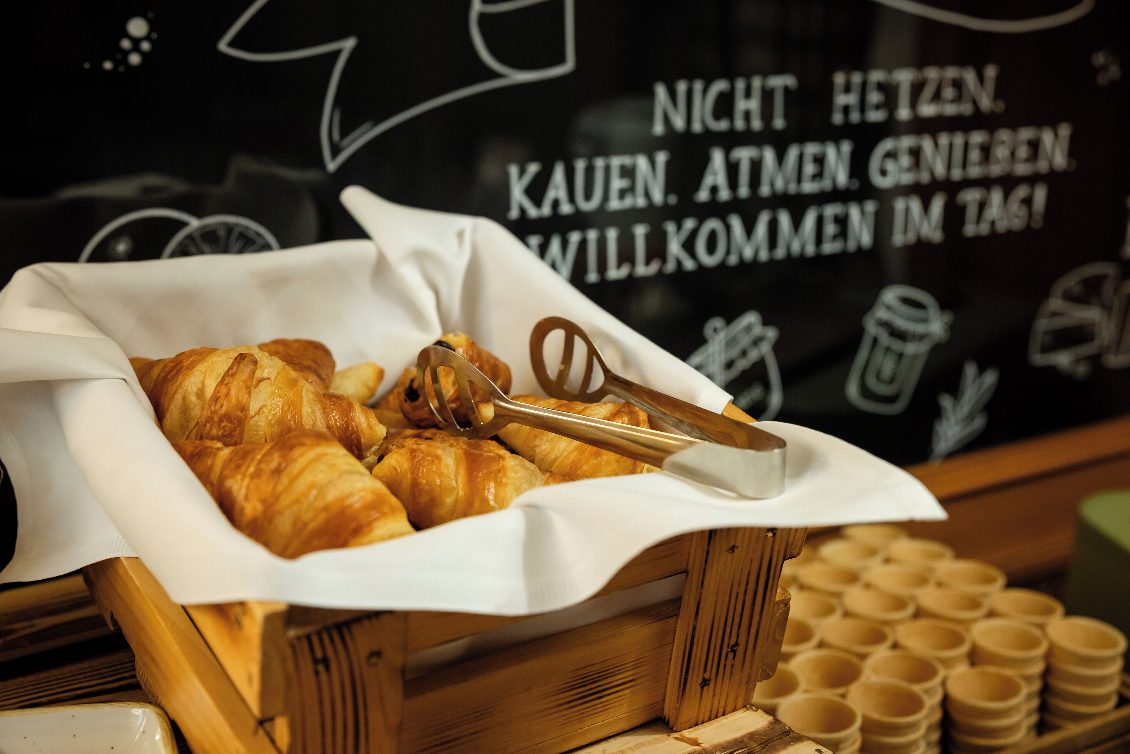 A wooden basket filled with croissants and a pair of tongs, placed in front of a blackboard with white text.