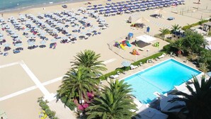 A beach with rows of umbrellas and sun loungers, a swimming pool, and palm trees.