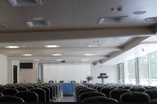 Un salón de conferencias con filas de sillas, una pantalla de proyector y un atril.