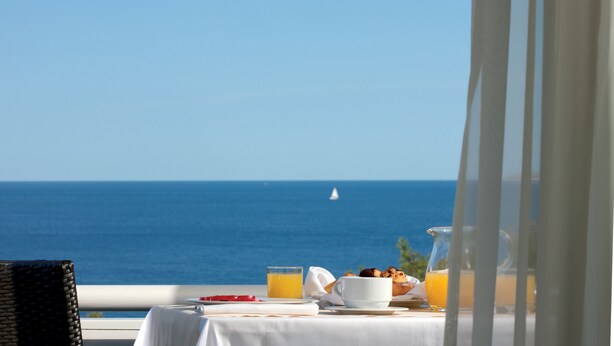 A table with breakfast items and a glass of orange juice, overlooking the sea.