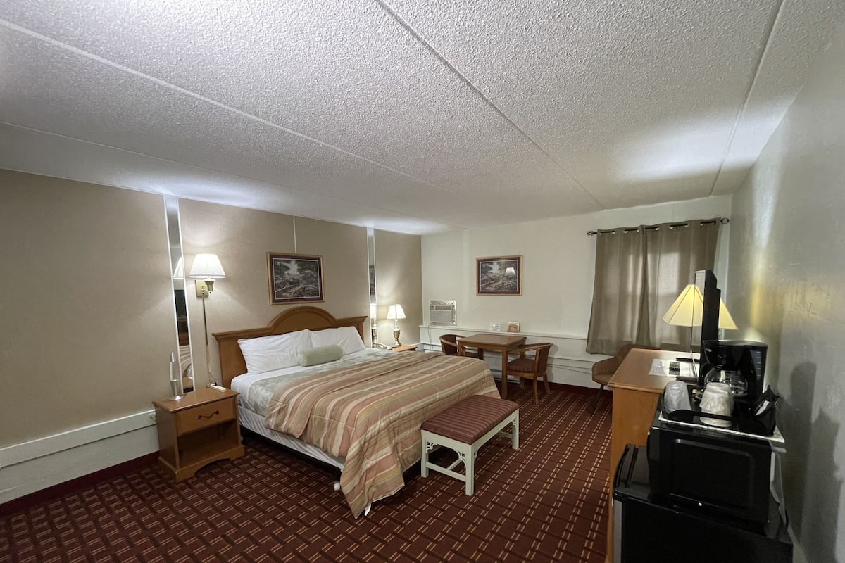 Habitación de hotel con una cama grande, un escritorio, una silla, una mesita, una lámpara y una cafetera.
